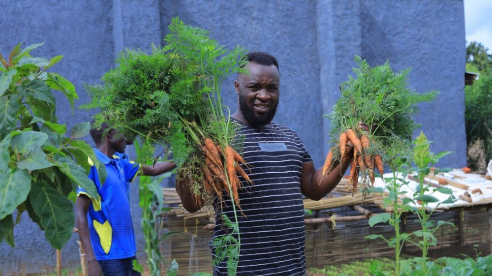 man holding carrots, showing permaculture harvest in Nakivale refugee settlement, Uganda (Unidos)